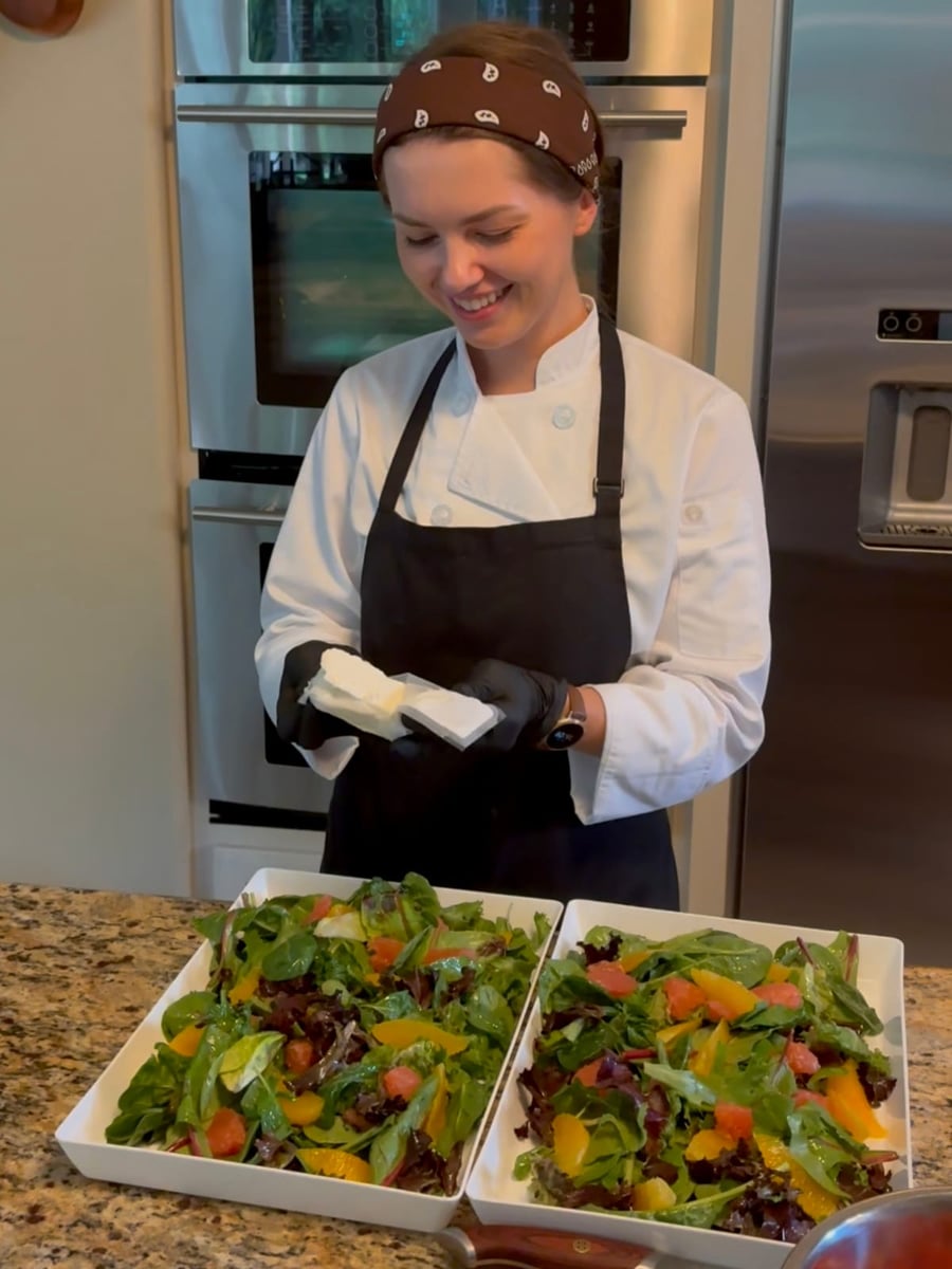Chef Ana crumbling honey goat cheese over two trays of fresh citrus salad with spring mix, grapefruit and orange segments.