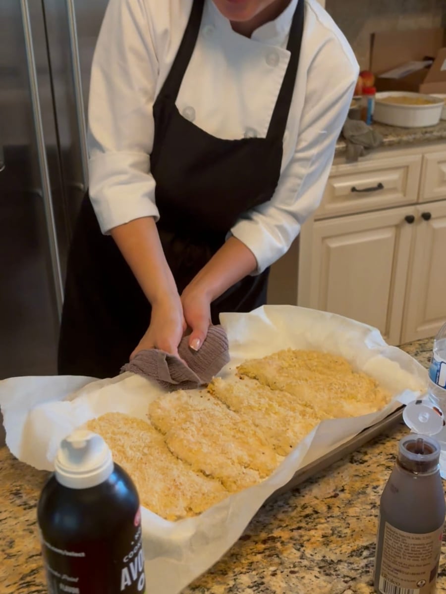 Chef Ana pressing a crisp panko crust onto chicken cutlets for the baked Chicken Milanese on a parchment-lined tray.