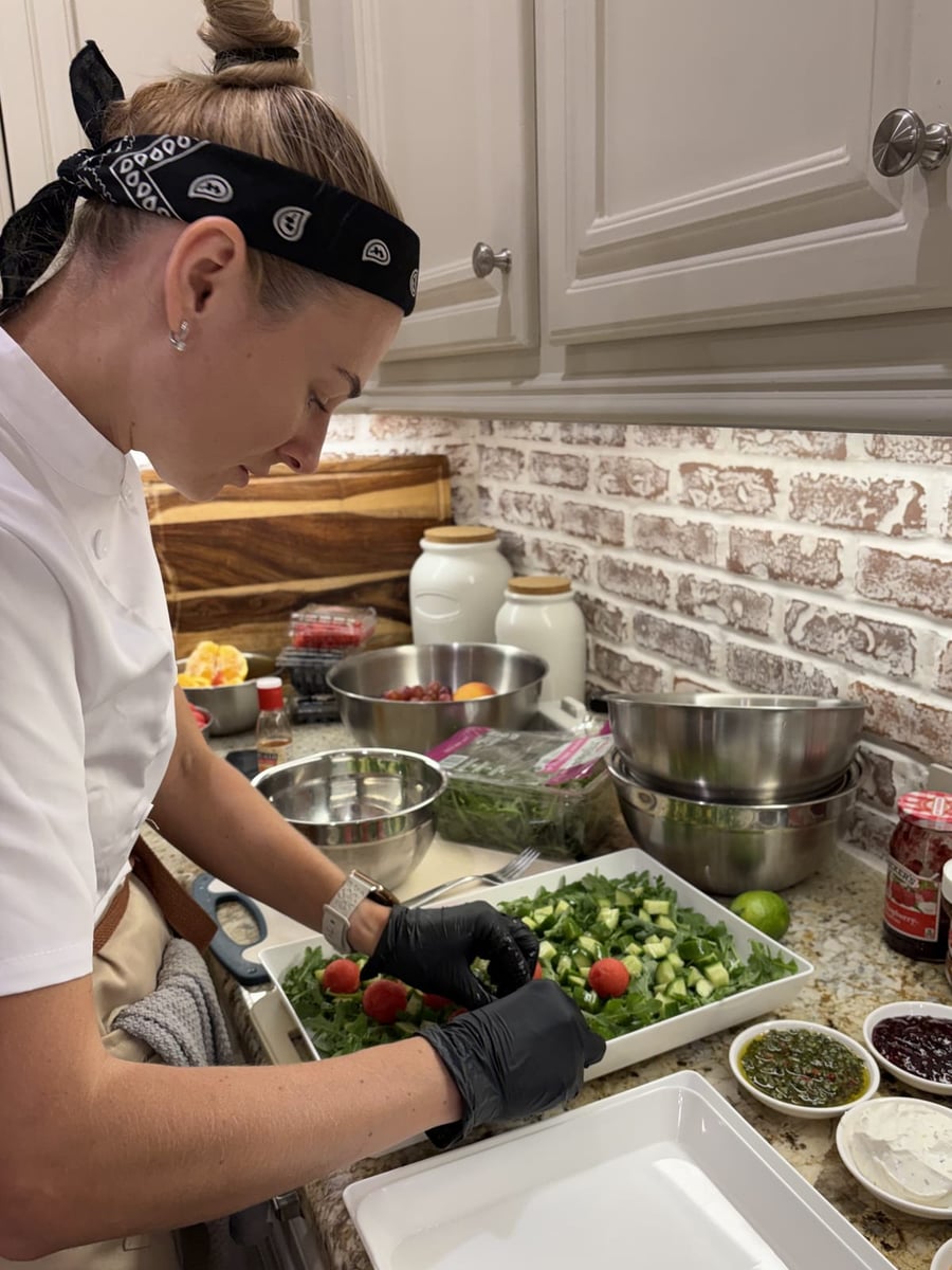 Chef Ana plating a fresh summer salad of arugula, cucumber and watermelon, with berries, jam and herb dressing set out on the counter.