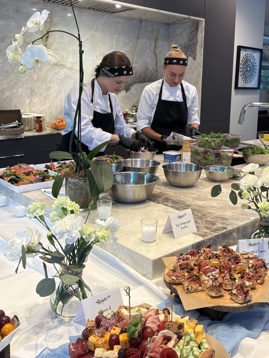 Chef Ana and her assistant tossing citrus salads at the marble island beside fig & gorgonzola crostini, prosciutto & caper crostini and a charcuterie board.