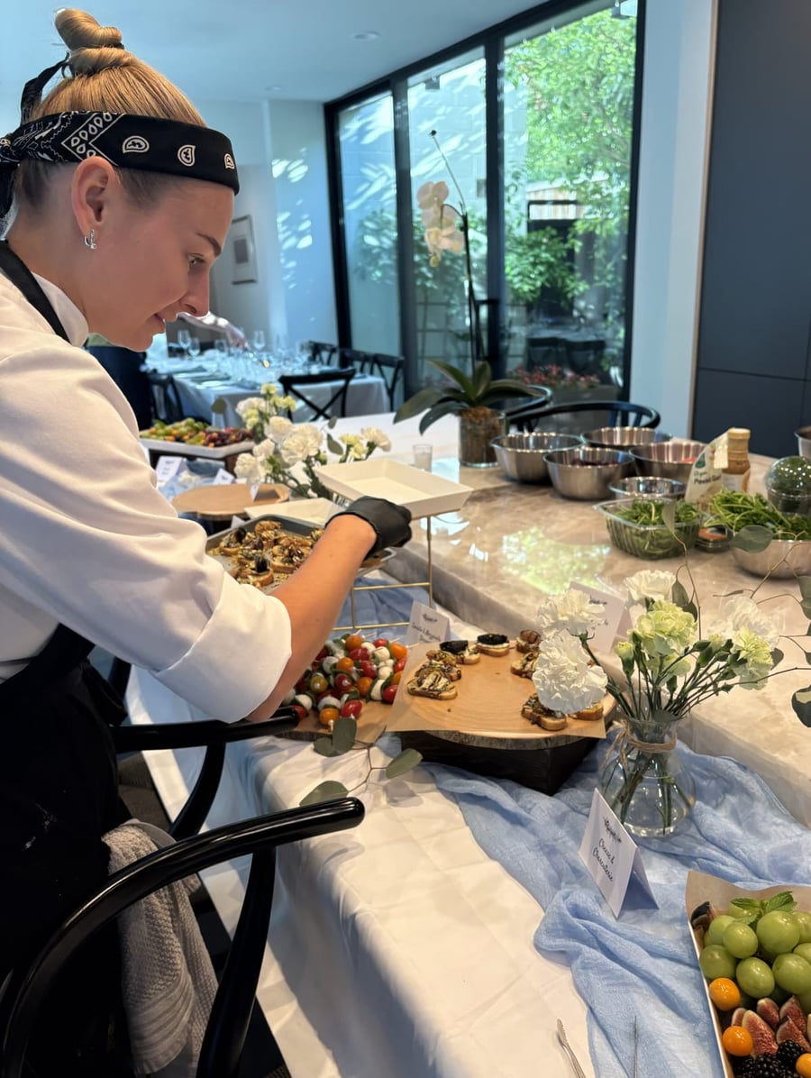 Chef Ana arranging the appetizer table — caprese skewers and crostini on a wood board — with the dining room set behind.