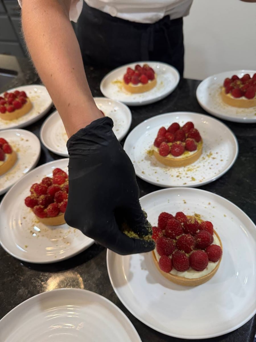 A gloved hand dusting crushed pistachio over individual raspberry tarts with vanilla cream on white plates.