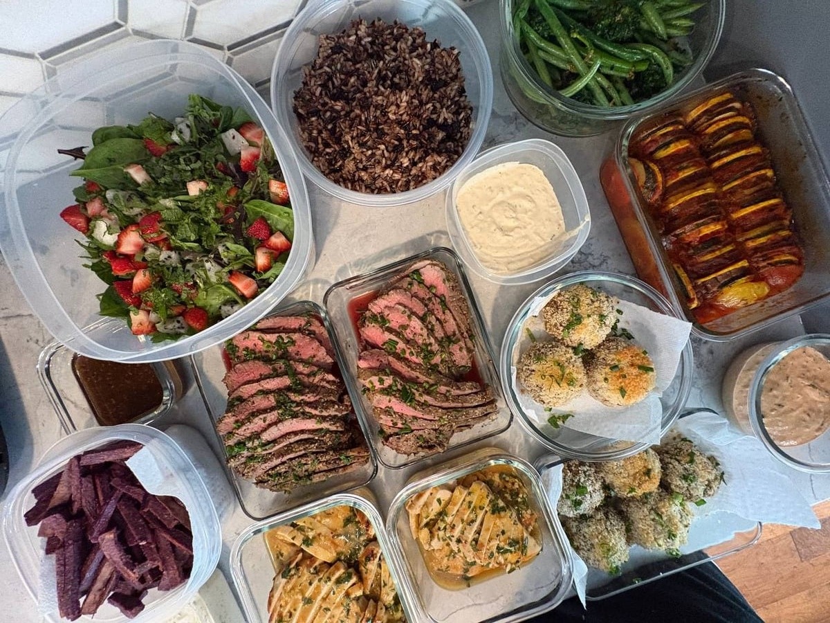 A collection of glass and plastic containers featuring sliced beef roast, orange chicken, crab cakes, ratatouille, purple sweet potato fries, and a fresh salad.