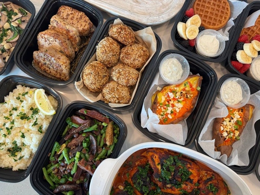 A variety of gourmet meal prep containers featuring herb-baked chicken, honey dijon pork chops, crab cakes, and fresh vegetable sides.