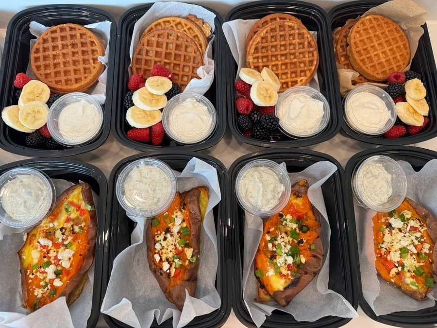 Nutritious breakfast meal prep containers featuring scrambled egg stuffed sweet potatoes with yogurt sauce and protein waffles with fresh berries.