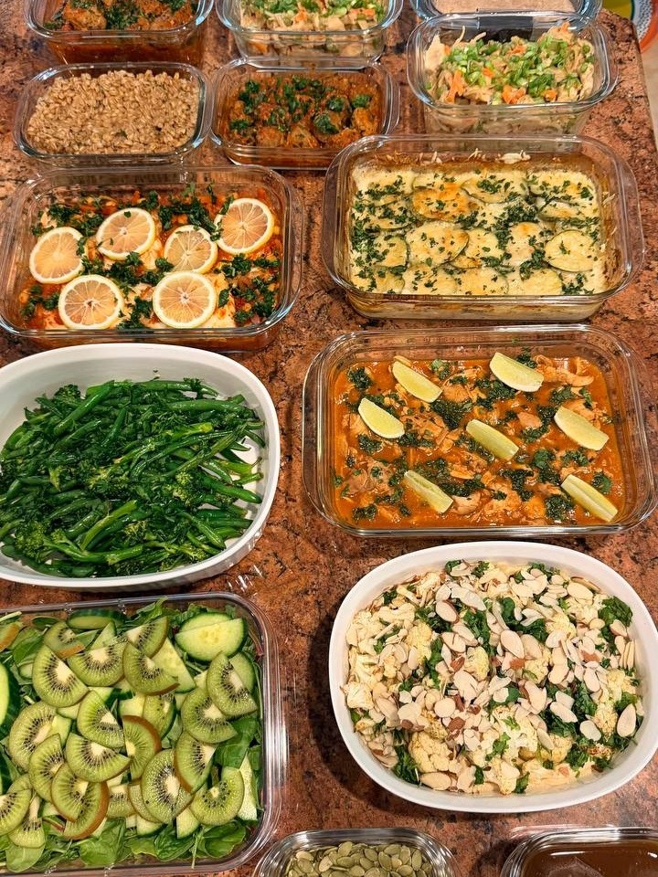 A variety of meal prep containers featuring saucy chicken, Tuscan meatballs, potato zucchini gratin, white fish, leafy salads, and roasted vegetables set on stone.