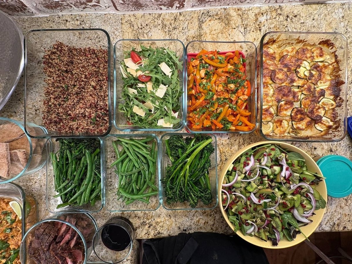 An assortment of glass meal prep containers featuring steak, herb-crusted chicken, buttery beans, roasted gratin, and a fresh kiwi-cucumber-spinach pumpkin seed salad.