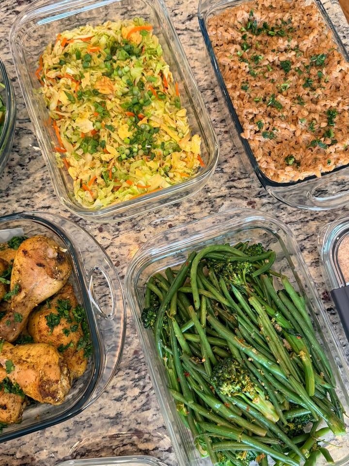 Glass containers holding Lebanese chicken drumsticks, herbed Italian farro, cabbage and carrot stir-fry, and chili crisp French beans and broccolini on a marble surface.