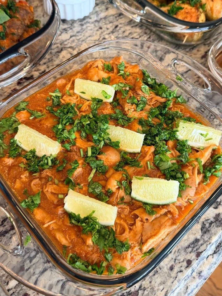 A glass baking dish filled with saucy shredded chicken thighs, topped with fresh chopped cilantro and lime wedges, presented on a granite countertop.