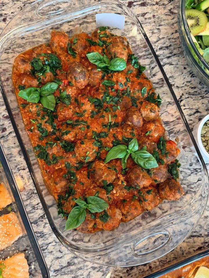 A glass baking dish filled with Tuscan-style beef meatballs poached in a vibrant tomato basil sauce, garnished with fresh whole basil leaves and chopped herbs.