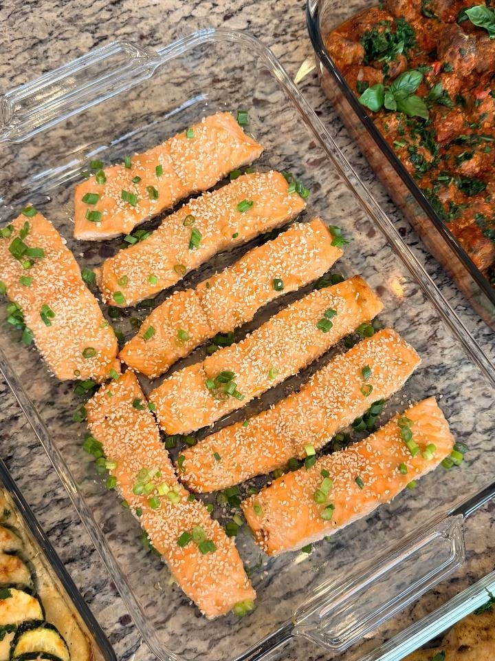 Top-down view of clear glass containers featuring miso-glazed salmon fillets topped with sesame seeds and savory Tuscan-style beef meatballs in rich tomato basil sauce.