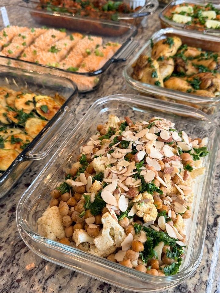 A glass container features Mediterranean spiced cauliflower, chickpeas, and fresh greens, generously topped with sliced almonds and aromatic herbs for a beautiful presentation.