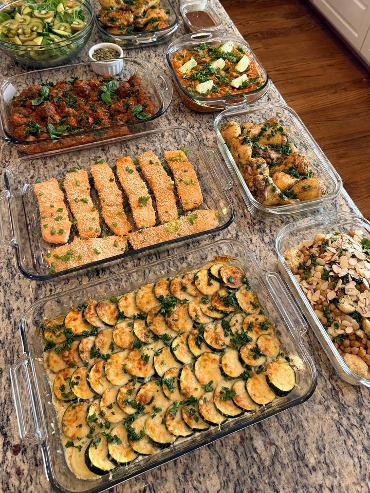 A wide spread of various glass containers filled with miso glazed salmon, Tuscan meatballs, chicken, vegetable gratin, and fresh salad on a kitchen counter.