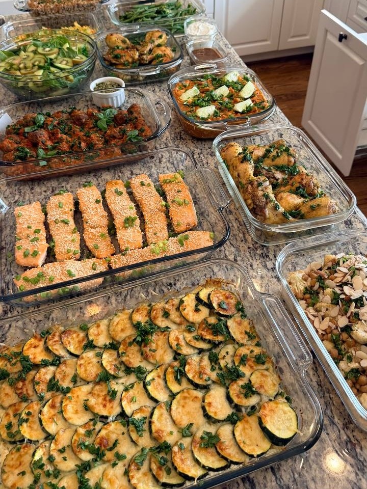 A variety of glass containers holding miso salmon, Tuscan meatballs, zucchini gratin, chicken, and salads, displayed attractively on a kitchen countertop for meal prep.