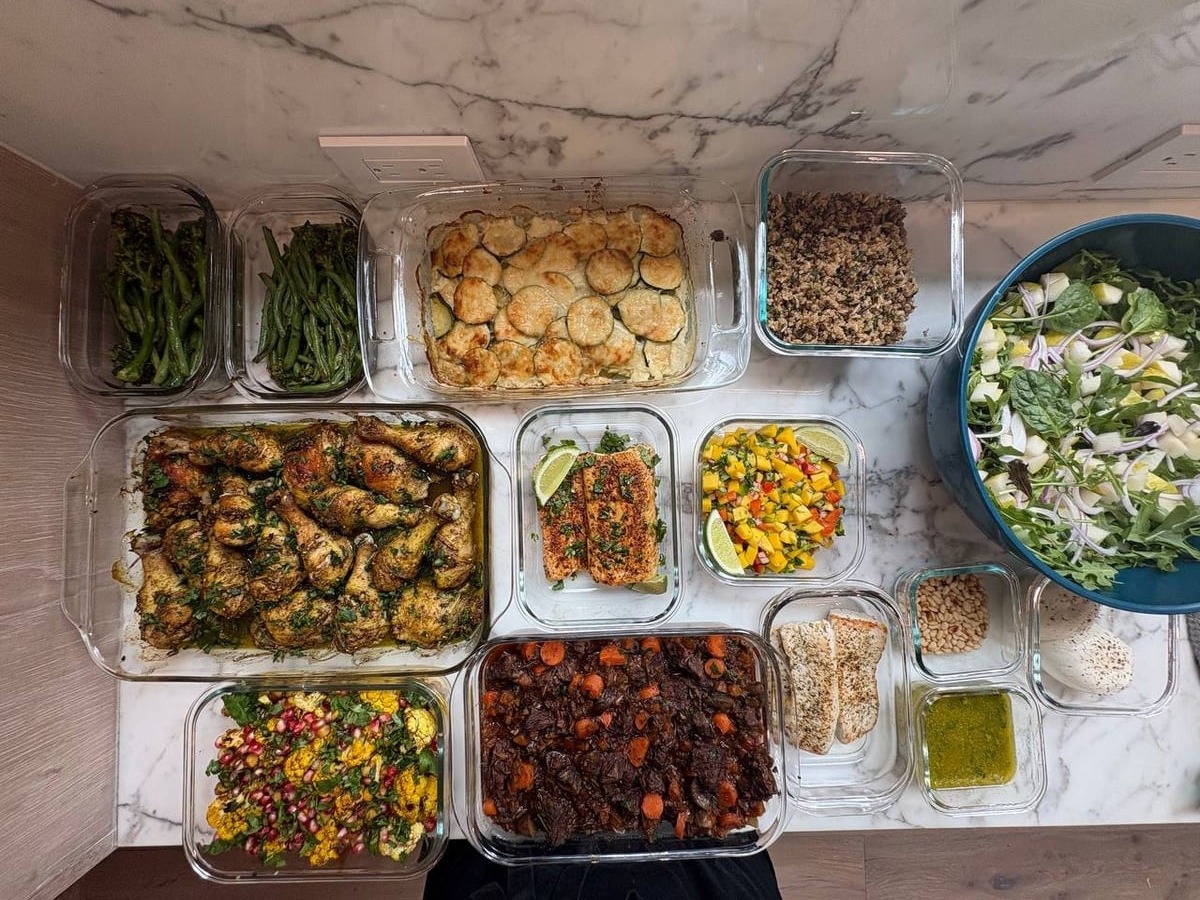 Top-down view of a sophisticated weekly meal prep service selection featuring zaatar chicken, potato gratin, and seasonal sides in clear glass containers.