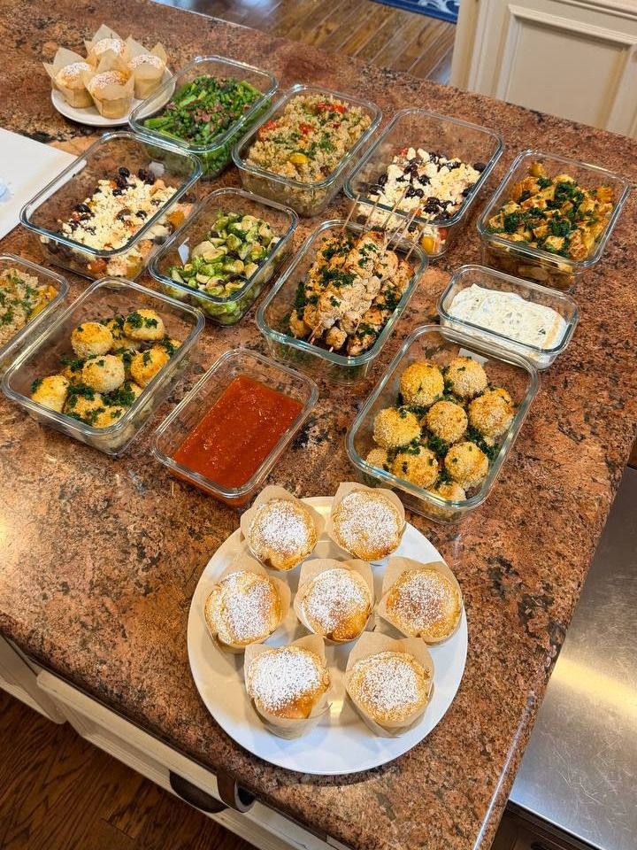 An overhead view of glass meal prep containers featuring chicken souvlaki, salmon bites, meatballs, vibrant grain salads, and raspberry muffins on a granite countertop.
