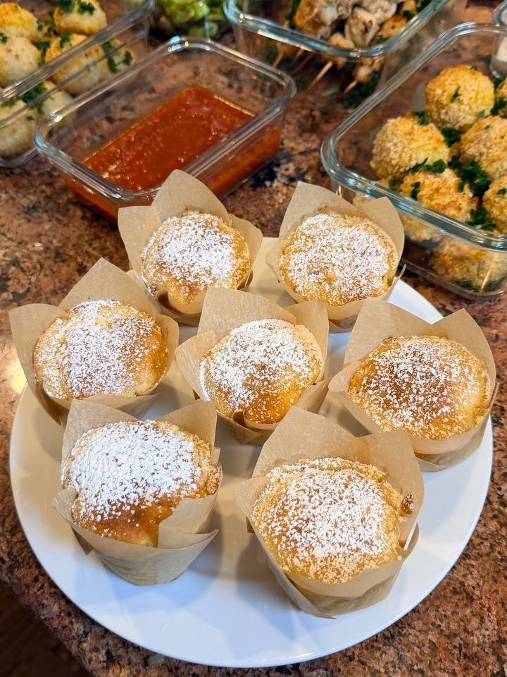 A white plate topped with raspberry white chocolate muffins next to glass containers filled with breaded mozzarella meatballs and chicken souvlaki skewers.