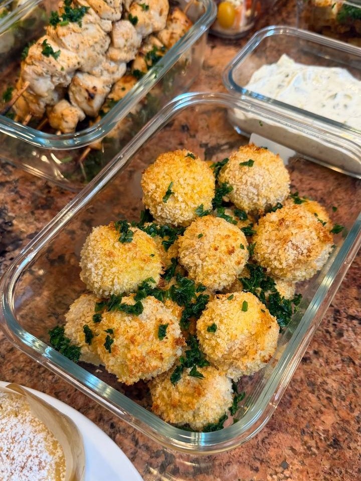 A glass meal prep container filled with crispy golden brown Kiev mozzarella meatballs garnished with fresh green herbs, displayed on a marble countertop.