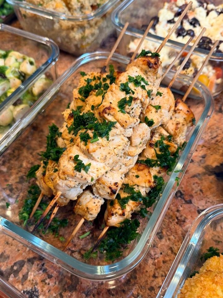 A glass container holds four Greek style chicken souvlaki skewers seasoned with herbs, presented alongside containers of quinoa salad and Mediterranean salmon.