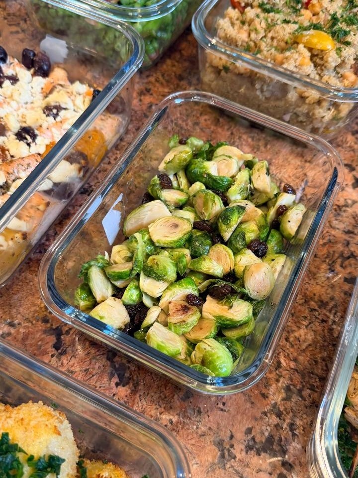 Glass meal prep containers filled with maple brussels sprouts, seared asparagus with pancetta, quinoa salad, mediterranean salmon bites, and breaded kiev mozzarella meatballs.