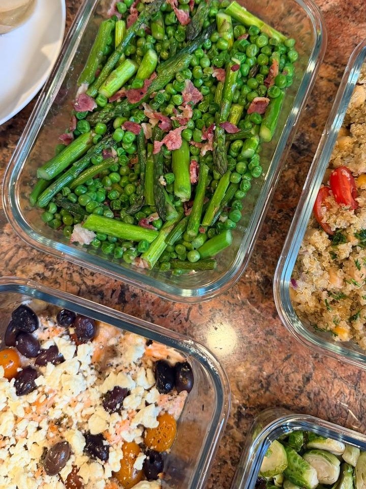 A glass container with seared asparagus, peas, and pancetta sits beside baked salmon with feta, olives, quinoa salad, and a raspberry white chocolate muffin.