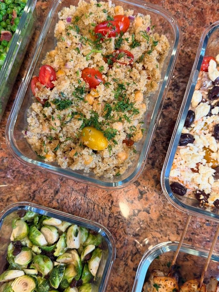 Glass meal prep containers filled with quinoa chickpea salad, Mediterranean salmon bites with olives, and herb-grilled chicken skewers next to roasted Brussels sprouts.