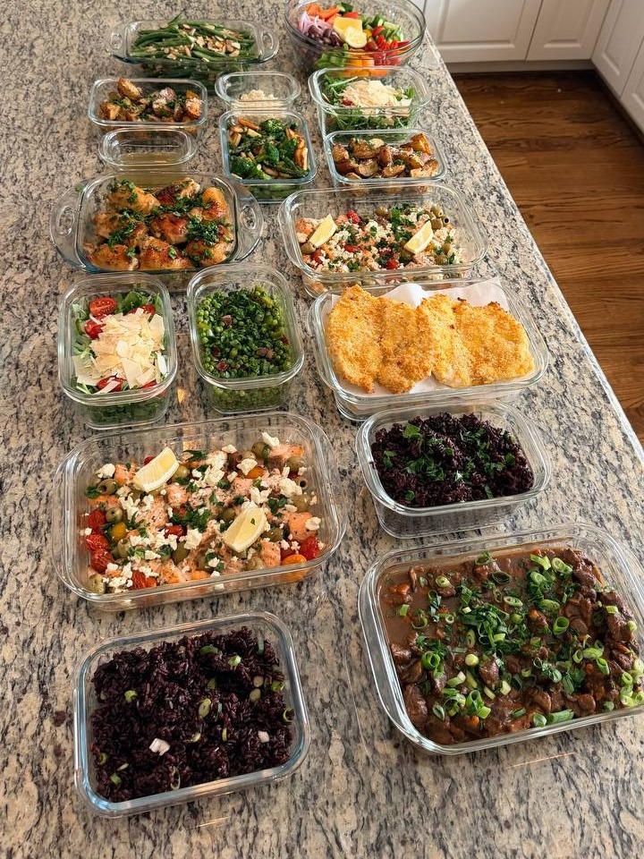 A variety of glass containers filled with beef burgundy, baked salmon, chicken milano, and various side dishes neatly arranged on a granite kitchen island.