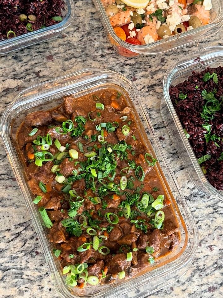 Glass meal prep containers filled with rich beef burgundy stew and separate portions of black rice topped with fresh scallions and herbs.