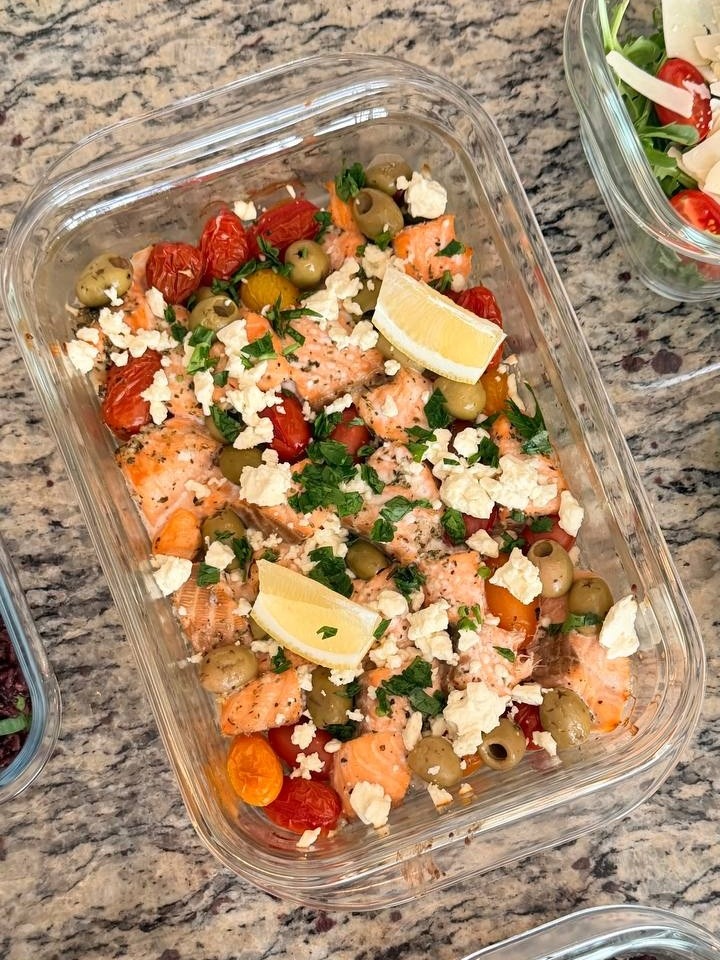 Glass container filled with Mediterranean baked salmon bites topped with green olives, crumbled feta cheese, fresh herbs, and bright lemon wedges for garnish.