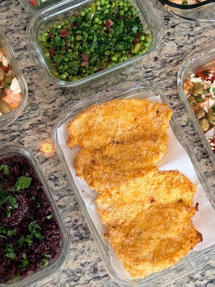 Glass meal prep containers filled with breaded chicken cutlets, black rice, seared bell peppers and peas, and a fresh arugula salad with parmesan.