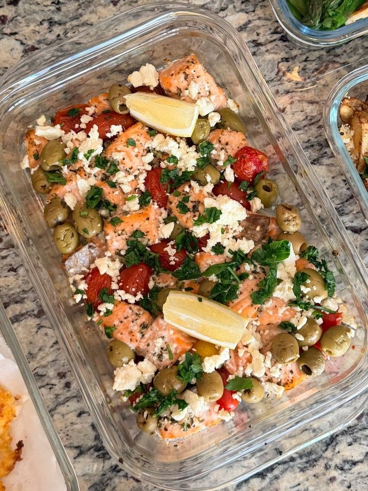 A glass container holds Mediterranean baked salmon bites topped with crumbled feta cheese, green olives, cherry tomatoes, fresh herbs, and lemon wedges.