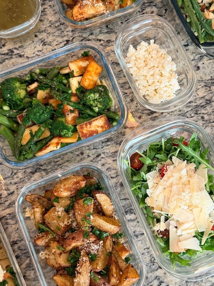 Glass containers featuring truffle parmesan baby potatoes, sautéed broccoli with paneer, and an arugula salad topped with shaved parmesan and fresh tomatoes.