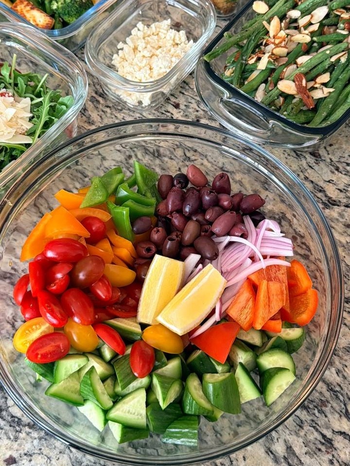 Glass containers holding classic Greek salad, green beans almondine, truffle parmesan baby potatoes, sauteed broccoli with paneer, and a bowl of fresh feta cheese.