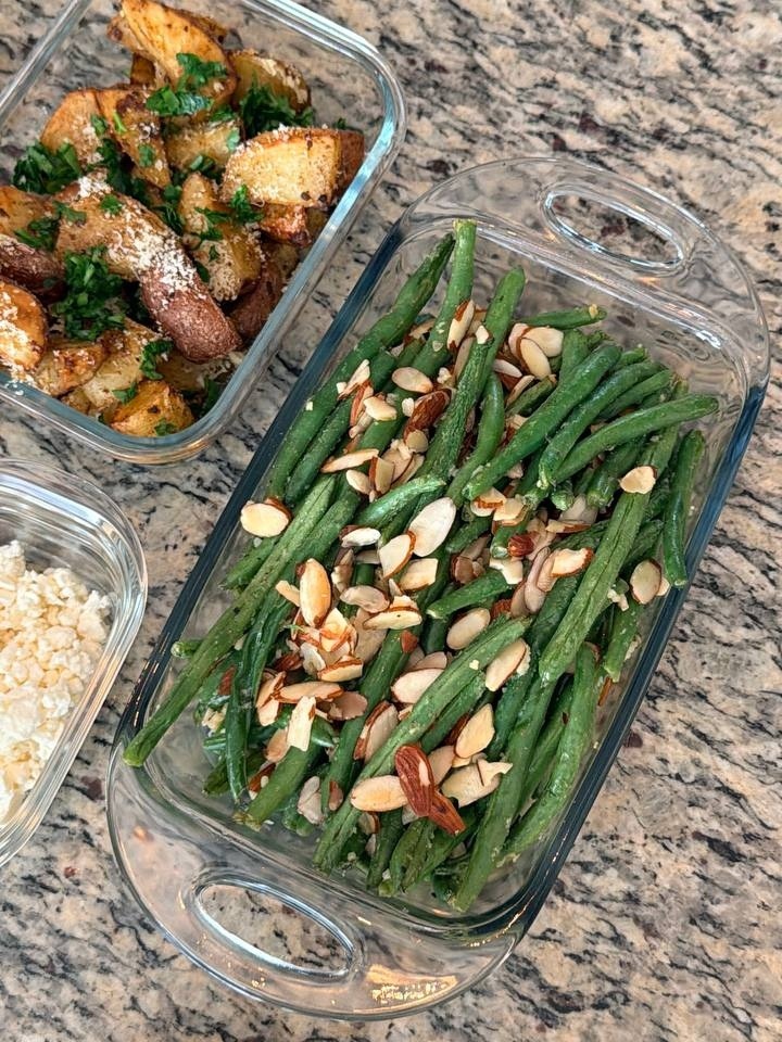 Glass containers holding roasted truffle parmesan baby potatoes and fresh green beans almondine with toasted almond slivers, arranged nicely on a granite countertop.