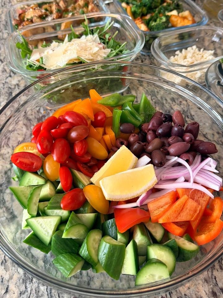A large glass bowl filled with fresh Greek salad ingredients including cherry tomatoes, cucumber slices, kalamata olives, bell peppers, red onions, and lemon wedges.
