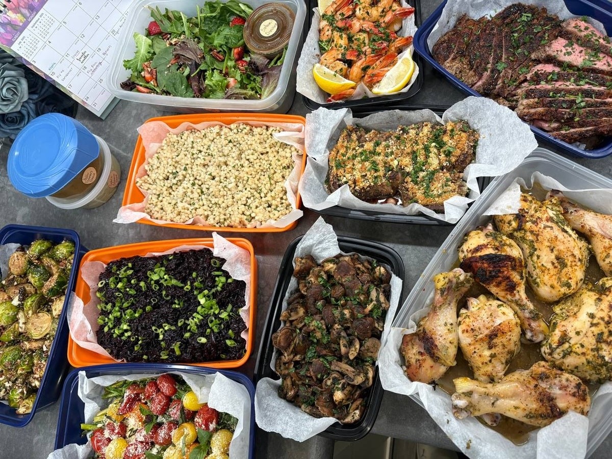 An overhead shot of various meal prep containers featuring orange ginger roasted chicken, herb crusted steak, garlic shrimp, black rice, and cranberry brussel sprouts.