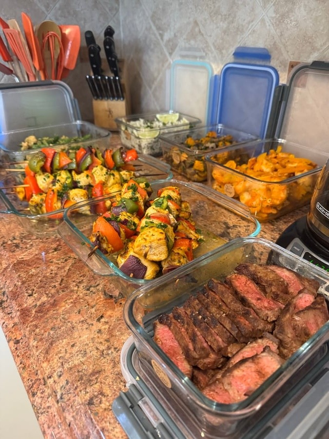 A variety of glass containers filled with chicken skewers, sliced NY strip steak, shrimp fajitas, and side dishes arranged for meal preparation.