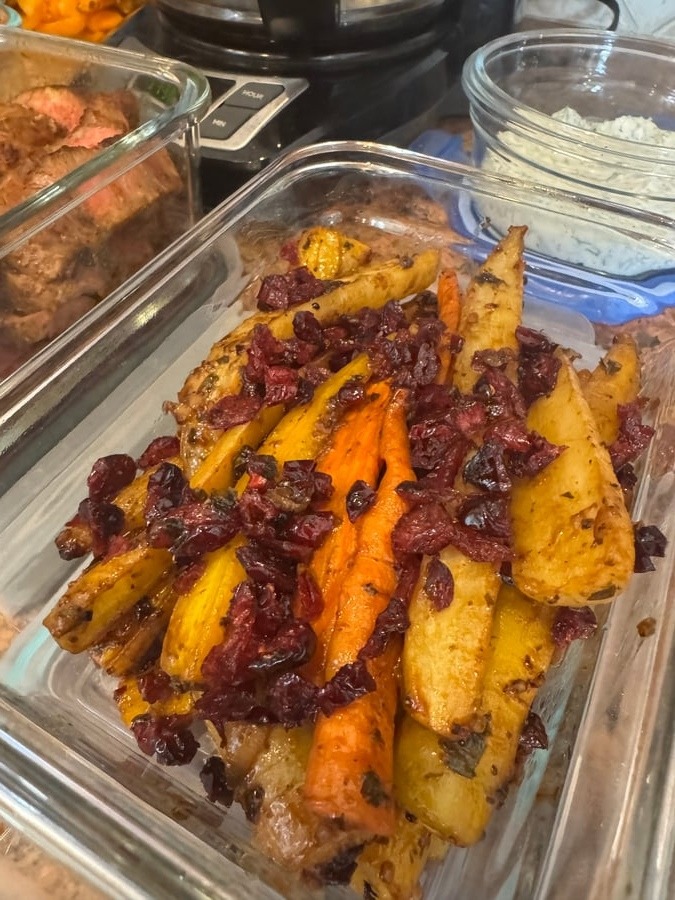 A glass container filled with balsamic cranberry baked carrots, featuring golden roasted root vegetables topped with dark, sweet dried cranberries and savory herbs.