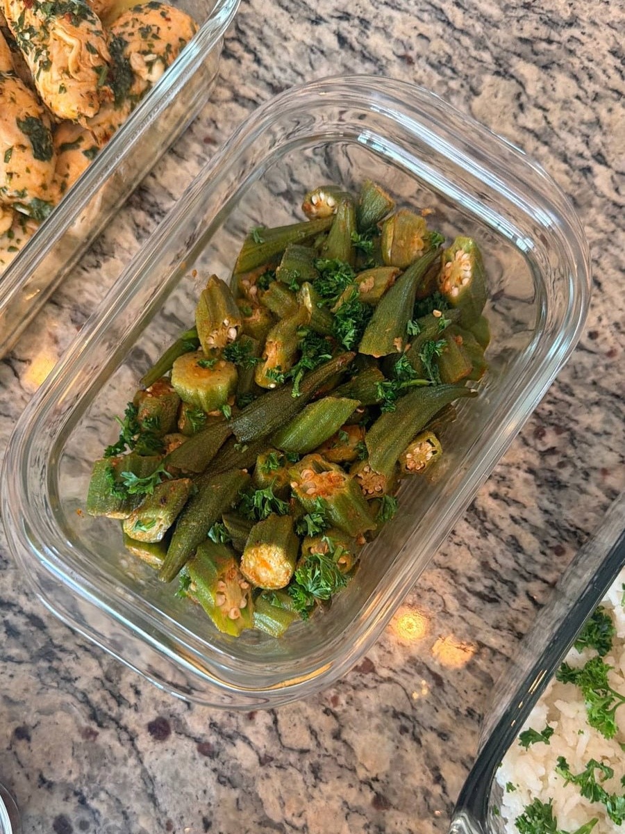 A glass storage container filled with freshly roasted garlic paprika baked okra, garnished with bright green parsley, resting on a speckled granite countertop.