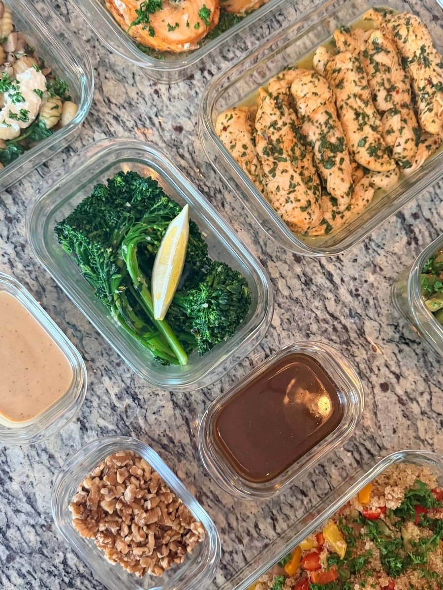 Glass containers displaying spinach stuffed salmon pinwheels, honey lime chicken tenders, lemon garlic broccolini, roasted peppers quinoa, and toasted walnuts on stone surface.