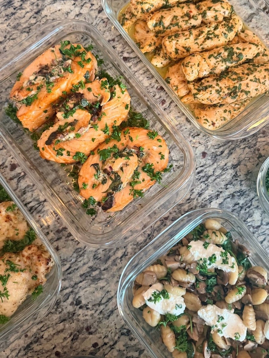 Glass meal prep containers feature spinach-stuffed salmon pinwheels, honey lime chicken tenders, and mushroom spinach gnocchi topped with creamy fresh burrata cheese in sauce.