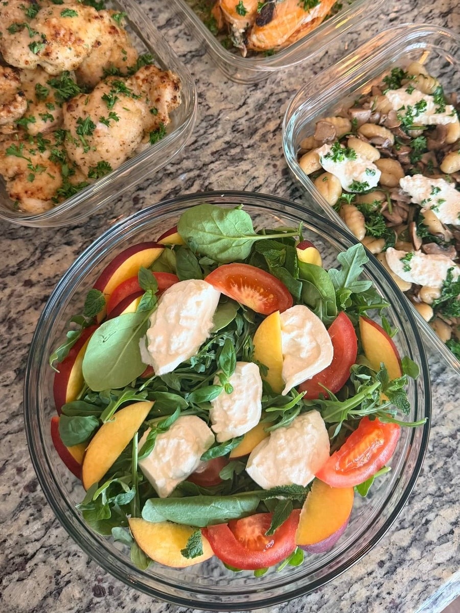 Glass meal prep containers filled with spinach stuffed salmon, garlic parmesan chicken, mushroom gnocchi with burrata, and a fresh peach mozzarella salad.