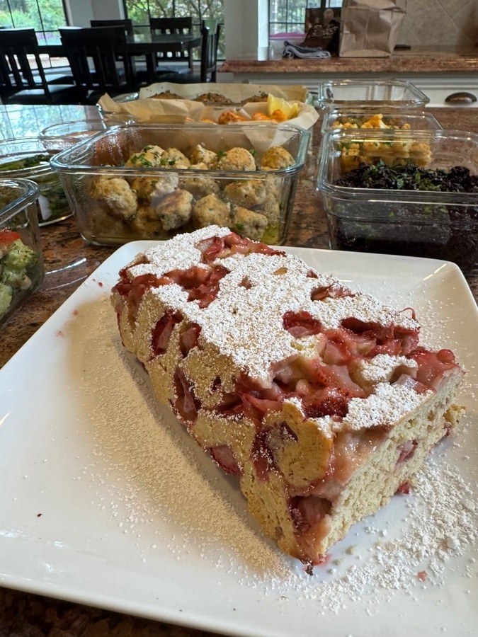 A golden-brown gluten-free strawberry shortcake loaf dusted with powdered sugar, sitting on a square white plate, with meal prep containers in the background.