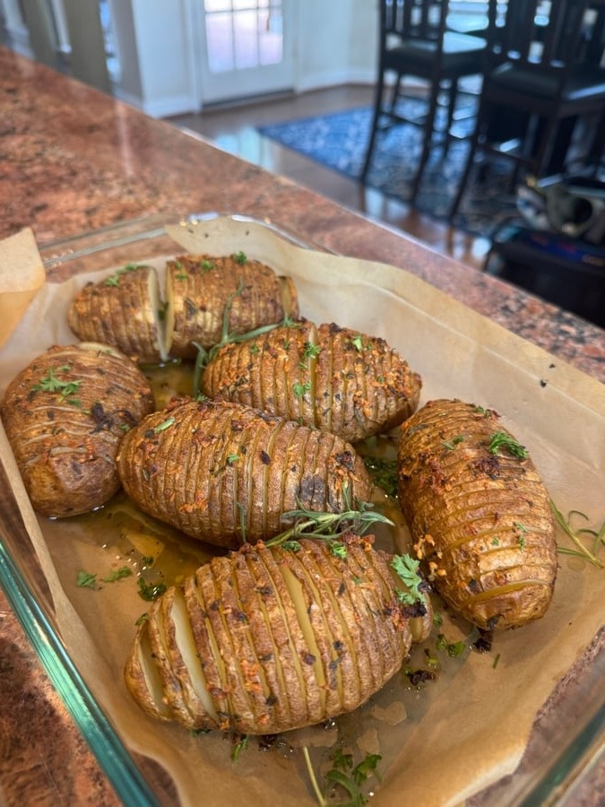 A glass baking dish lined with parchment paper holds several oven-roasted hasselback potatoes seasoned with fresh rosemary, garlic, and bright green parsley.