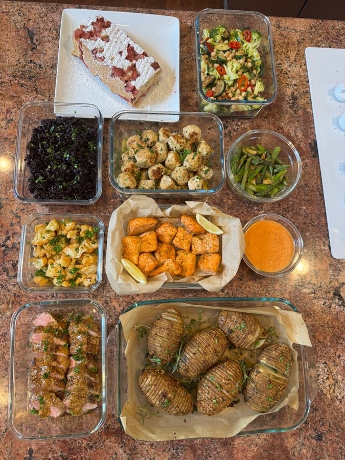 An overhead view of varied meal prep containers featuring garlic salmon, chicken meatballs, pork tenderloin, black rice, roasted potatoes, asparagus, cauliflower, and strawberry loaf.