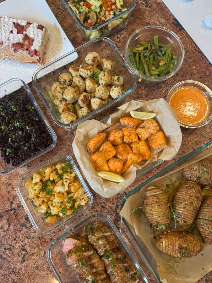 A variety of glass meal prep containers featuring garlic salmon bites, chicken picatta meatballs, pork tenderloin, husselback potatoes, stir-fry vegetables, and a strawberry loaf.