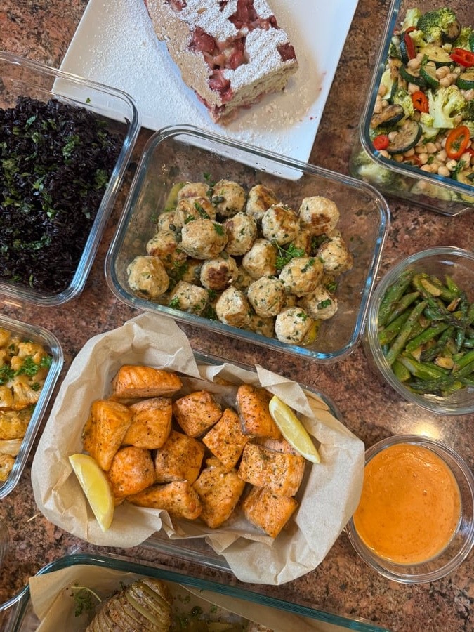 A glass container display of garlic salmon bites, chicken picatta meatballs, cilantro black rice, chili seared broccoli, roasted asparagus, and strawberry shortcake.