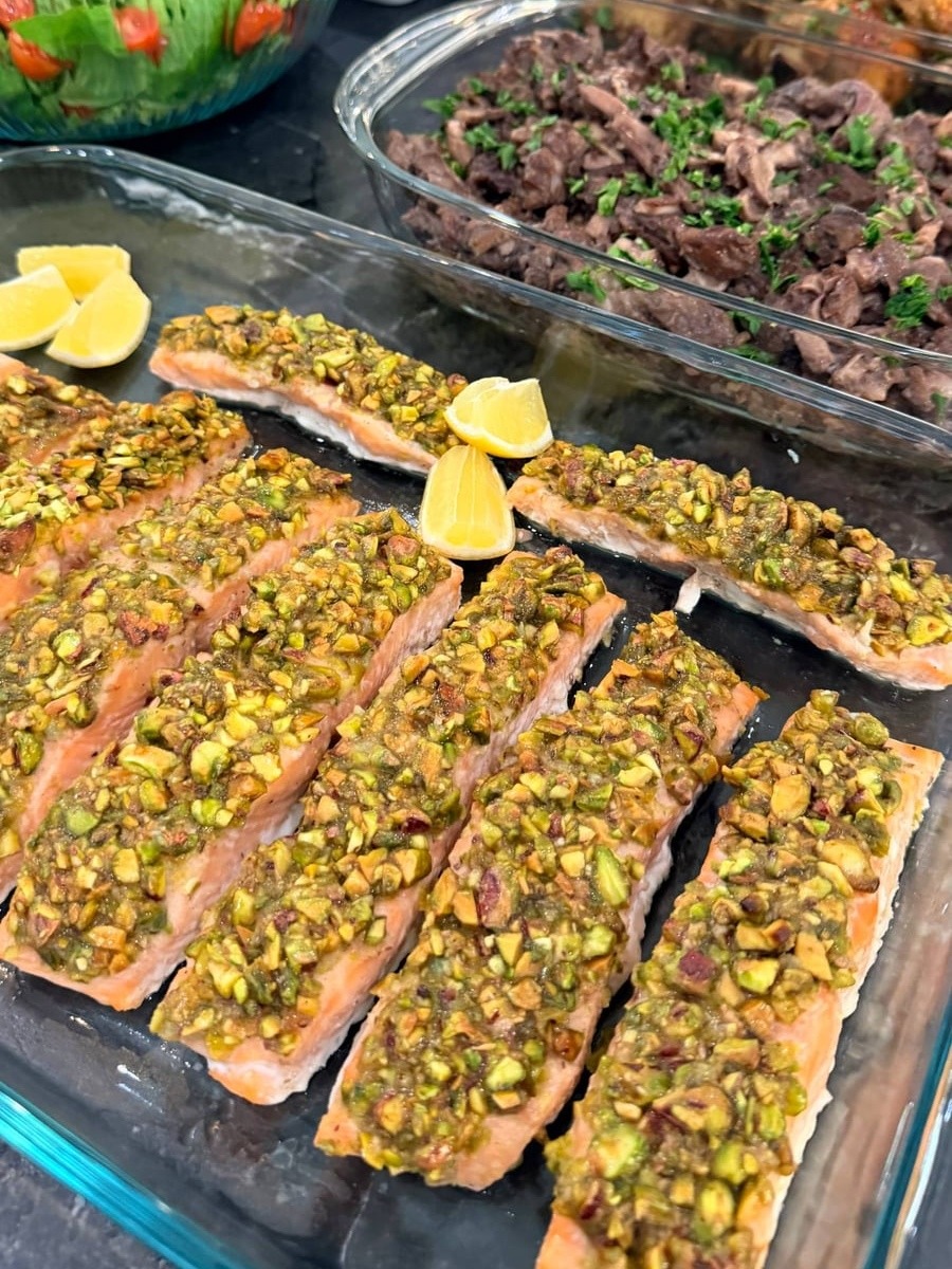 A glass tray filled with oven-baked pistachio-crusted salmon fillets garnished with fresh lemon wedges, alongside various sides in clear serving containers.