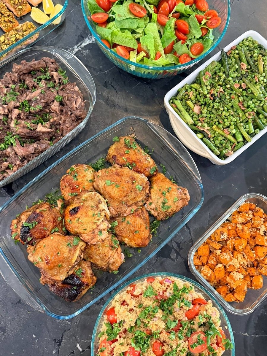 A selection of glass containers showcasing pistachio salmon, balsamic chicken, seared asparagus, quinoa salad, beef ribeye, and a fresh Caesar salad.