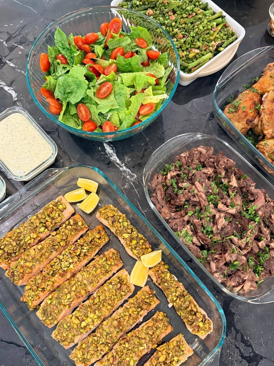 A selection of prepared dishes including pistachio crusted salmon, sauteed beef ribeye with mushrooms, Caesar salad, and seared asparagus served in glass containers.